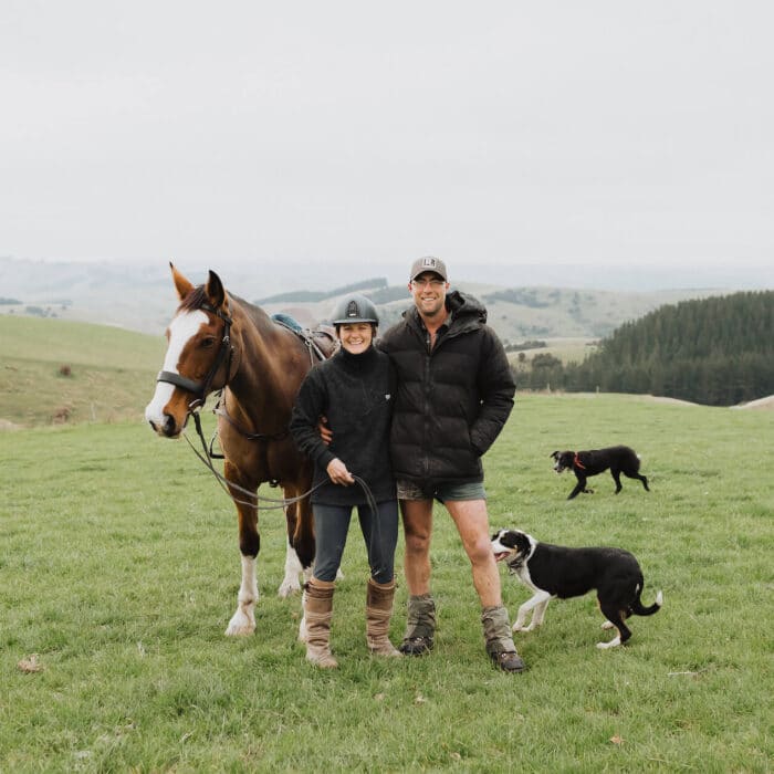 Woman and man stand with their horse and two farm dogs on a big grassy hill