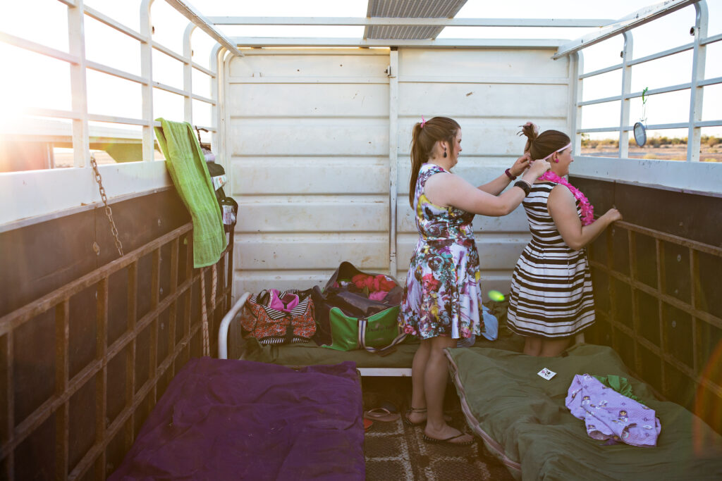Woman helps another woman do up the back of her dress, in the back of an open top truck trailer, in the outback.
