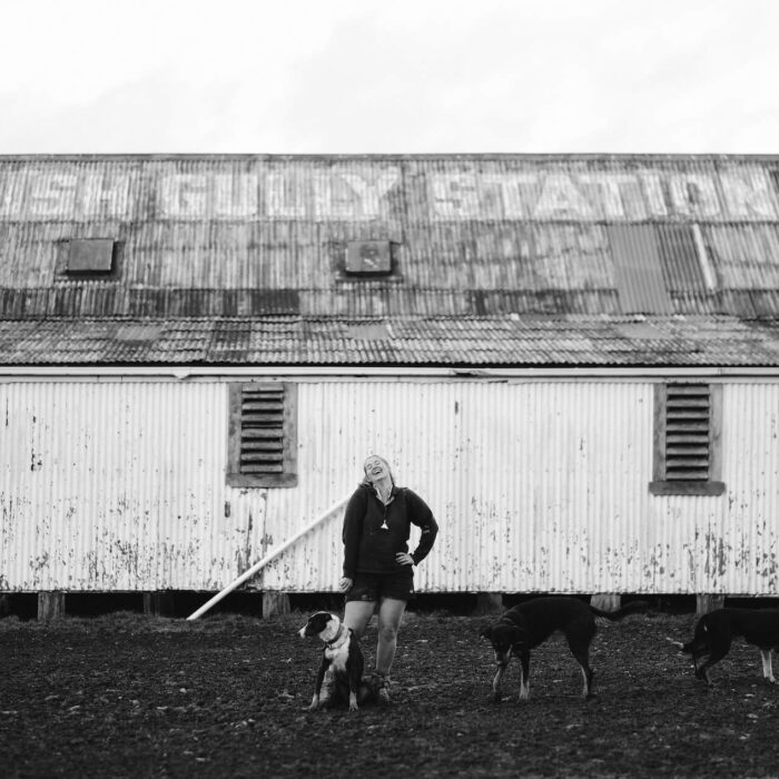 Black and white image of a young female shepherd standing in front of a large barn with two dogs.